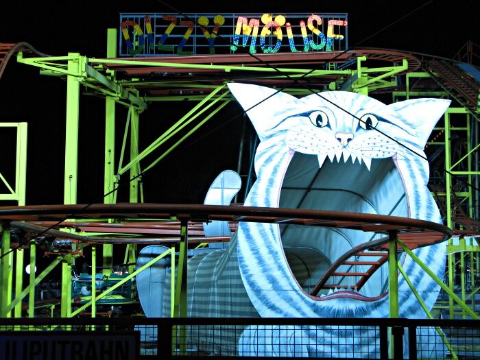A night-time color photo of a roller coaster, with the track going into the mouth of a giant cat, and a sign saying "Dizzy Mouse"